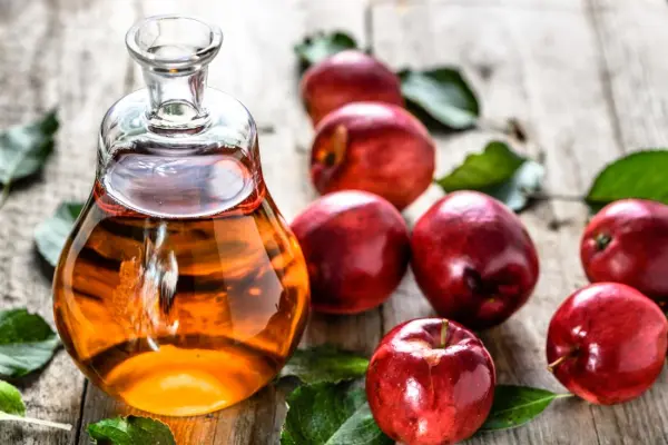 Bình thủy tinh chứa giấm táo và vài quả táo đỏ xung quanh nó trên nền gỗ.